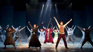 Ryhmä värikkäisiin aikakauden pukuihin pukeutuneita esiintyjiä seisoo lavalla leveässä V-muodostelmassa kädet iloisesti kohotettuna kirkkaiden näyttämövalojen ja tähtitaustan alla.