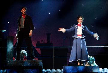 Kaksi esiintyjää seisoo yöllistä kattoa muistuttavalla lavasteella, jonka taustalla on savupiipun siluetteja ja tähtitaivas. Toisella on tumma asu ja lippis, toisella sininen hameasu, punainen rusetti ja valkoiset käsineet, kädet ojennettuina.