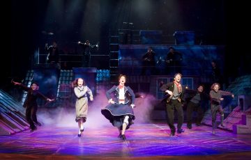 Vilkkaassa näyttämöesityksessä aikakauden pukuihin pukeutuneet näyttelijät tanssivat tarmokkaasti dramaattisessa valaistuksessa, ja taustalla on katto ja näyttelijäkaarti.