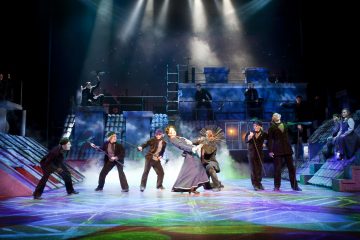 Näyttelijät esittävät vilkkaan musiikkikohtauksen näyttämöllä, jossa on värikäs valaistus, sumuefektejä ja kattojen tausta tähtitaivaan alla; keskeiset hahmot tanssivat, kun muut katselevat tai poseeraavat dramaattisesti.