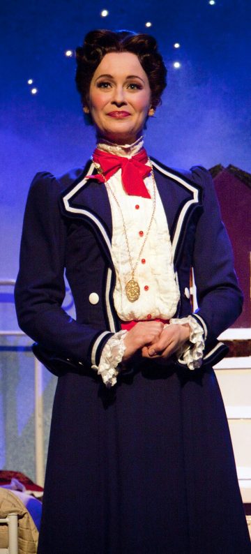 Mary Poppinsiksi pukeutunut nainen, jolla on sininen viktoriaaninen takki, jossa on valkoiset reunukset, punainen solmio ja kultainen riipus, seisoo lavalla sinisellä tähtitaustalla hymyillen ja kädet ristissä.