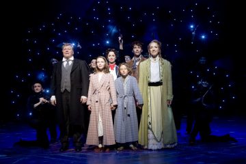 Ryhmä aikakauden pukuihin pukeutuneita näyttelijöitä seisoo rivissä lavalla, ja taustalla näkyy tähtimäisiä sinisiä valoja. Edessä seisoo kaksi kaapuihin pukeutunutta lasta, jotka pitävät aikuisia kädestä kiinni. Muut esiintyjät seisovat ja polvistuvat heidän takanaan.