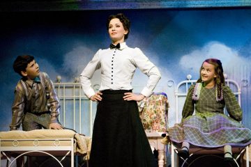 Valkoiseen puseroon ja mustaan hameeseen pukeutunut nainen seisoo itsevarmasti kahden erillisillä sängyillä istuvan lapsen välissä, ja sininen tausta viittaa oudon tai teatterinomaiseen ympäristöön. Lapset katsovat naista ihaillen.