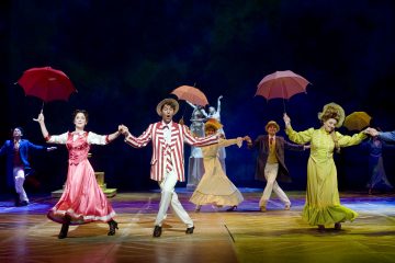 Värikkäisiin aikakauden pukuihin ja hattuihin pukeutuneet esiintyjät laulavat ja tanssivat lavalla pitelemässä punaisia ja vaaleanpunaisia sateenvarjoja teatterivalaistuksen alla, ja taustalla on maalattu tausta ja kaksi valkoista tanssijaa.