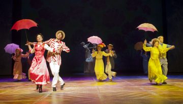 Esiintyjät lavalla pukeutuvat värikkäisiin vintage-pukuihin; kaksi etutanssijaa pukeutuu punaiseen ja valkoiseen, kun taas toiset keltaiset pitävät sateenvarjoja, mikä luo eloisan musiikkiteatterikohtauksen.