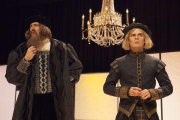 Lavalla seisoo kaksi hienostuneisiin renessanssiasuihin pukeutunutta miestä kattokruunun alla. Toisella on pitkä parta ja musta hattu, kun taas toinen on sileäpartainen ja vaaleahiuksinen, ja molemmat näyttävät olevan syvällä ajatuksissa tai keskustelussa.