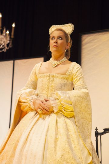Lavalla seisoo nainen, joka on pukeutunut hienostuneeseen kulta-valkoiseen aikakauden pukuun, jossa on helmiä ja koristeellinen päähine, ja näyttää yllättyneeltä tai säikähtäneeltä. Taustalla näkyy kattokruunu ja vaaleat paneelit.