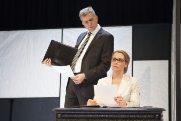 Pukumies seisoo minimalistisessa, modernissa näyttämöympäristössä, kädessään kansio ja papereita, ja katsoo alaspäin istuvaan, silmälasipäiseen ja valkoiseen bleiseriin pukeutuneeseen naiseen, jolla on myös papereita kädessään.