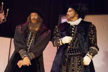 Lavalla seisoo kaksi miestä taidokkaissa renessanssipuvuissa. Toinen näyttää yllättyneeltä silmät suurina ja partaisena, kun taas toinen, jolla on höyhenpukuinen hattu ja koristeellinen tunika, katsoo häntä ankarasti. Taustalla roikkuu kattokruunu.