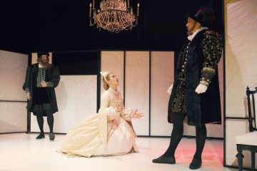 Nainen, jolla on koristeellinen kermanvärinen mekko, polvistuu lavalla ja osoittaa ylöspäin kohti miestä, jolla on hienostunut tumma puku. Taustalla seisoo toinen aikakauden vaatteisiin pukeutunut mies. Heidän yläpuolellaan roikkuu kattokruunu.