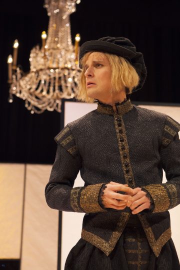 Mustankultaiseen renessanssipukuun ja -hattuun pukeutunut näyttelijä seisoo kattokruunun alla ja katsoo sivulle vakavalla ilmeellä.