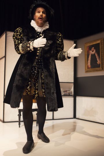 Henkilö, jolla on koristeellinen, renessanssityylinen puku, johon kuuluu röyhelökaulus, turkisverhoiltu takki ja valkoiset käsineet, elehtii dramaattisesti lavalla. Heidän takanaan seinällä roikkuu muotokuva.