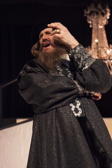 Koristeellisiin, tummiin renessanssityylisiin vaatteisiin ja tekopartaan pukeutunut henkilö nostaa dramaattisesti kätensä ja esiintyy lavalla, jonka taustalla näkyy kattokruunu.