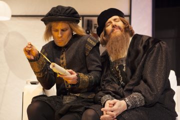 Kaksi miestä istuu tiiviisti vierekkäin taidokkaissa renessanssipuvuissa. Toinen tarkastelee kääröä keskittyneesti, kun taas toinen, jolla on pitkä tekoparta, sulkee silmänsä ja hymyilee tyynenä.