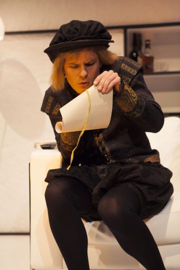 Historialliseen asuun pukeutunut henkilö istuu valkoisella tuolilla, pitää kädessään käärittyä paperia ja tutkii keskittyneenä kultaista narua. Taustalla näkyy hyllyjä, joissa on pulloja.