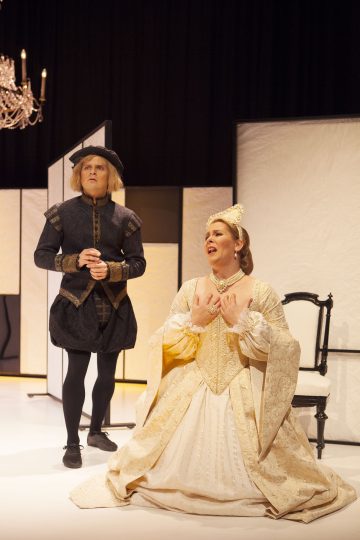 Tummiin renessanssityylisiin vaatteisiin pukeutunut mies seisoo koristeelliseen kermanväriseen ja kultaiseen pukuun pukeutuneen naisen vieressä, joka polvistuu ja laulaa dramaattisesti lavalla kattokruunun alla. Taustalla on minimaalinen, pehmeä valaistus.