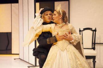 Tummiin renessanssipukuihin pukeutunut mies syleilee naista, jolla on koristeellinen, kulta-valkoinen ja kruunullinen puku, ja he katsovat toisiaan näyttämöllä dramaattisin ilmein. Taustalla on mustavalkoinen tuoli.