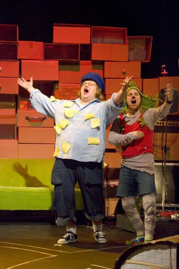 Kaksi ihmistä esiintyy näyttämöllä teatterillisesti; toisella on sininen hattu, sininen paita, joka on täynnä tarralappuja, ja tummat shortsit, kädet ojennettuina. Toisella on päällään lehtimäiset asusteet ja harmaat shortsit. Taustalla on värikkäitä laatikoita.