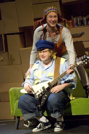 Kaksi ihmistä lavalla, toinen istuu vihreällä sohvalla ja soittaa sähkökitaraa, hänellä on sininen hattu ja silmälasit, vaatteissaan tarralappuja. Toinen seisoo takana hymyillen, yllään värikäs hattu ja haalarit.