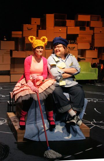 Kaksi ihmistä istuu lavalla, jonka lavasteena on pinottuja pahvilaatikoita. Toisella on vaaleanpunainen mekko ja oranssi eläinkorvallinen hattu, ja hänellä on kädessään moppi. Toisella on farkut, lenkkarit, sininen hattu ja tarralapuilla päällystetty paita.