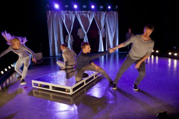 Neljä rennosti pukeutunutta henkilöä esittää dynaamisen tanssin lavalla, jossa on kirkkaat valot ja heijastava lattia. Yksi henkilö ojentaa kätensä auttaakseen toista ylös, kun taas kaksi muuta liikkuu tarmokkaasti taustalla. Verhot roikkuvat heidän takanaan.