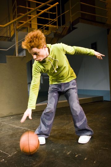 Henkilö, jolla on piikikäs punainen tukka, vihreä huppari ja leveälahkeiset farkut, dribblaa koripalloa sisäkentällä, jonka taustalla on portaita ja kaiteet.