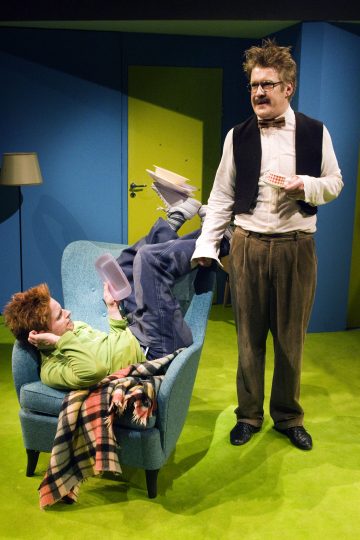 Henkilö makaa ylösalaisin sinisessä nojatuolissa lukemassa kirjaa, kun taas toinen henkilö, jolla on epäsiisti tukka ja silmälasit, seisoo lähellä ja pitää pientä tarjotinta. Kohtaus sijoittuu kirkkaanväriseen huoneeseen, jossa on vihreä matto.