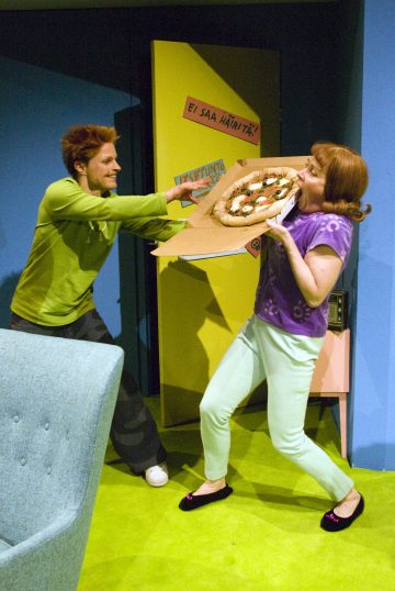 Vihreisiin pukeutunut henkilö työntää leikkisästi isoa pizzaa kohti toista pastellinväristä henkilöä, joka nojaa hämmästyneenä taaksepäin. He seisovat värikkäässä huoneessa, jossa on kirkkaat seinät ja huonekalut.