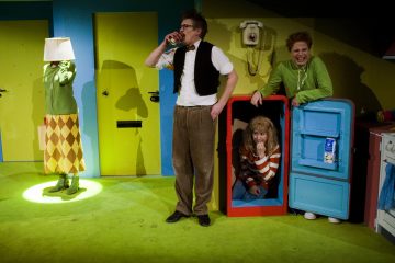 Neljä ihmistä värikkäässä huoneessa: yhdellä on lamppu päässään, yksi juo lasista, yksi hymyilee ja kyyristelee avoimen jääkaapin vieressä ja yksi istuu jääkaapin sisällä, kaikki omituisissa asuissa.