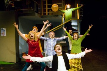 Seitsemän värikkäisiin retro-asuihin pukeutunutta aikuista seisoo lavalla kädet kohotettuna ja suu auki ja näyttää laulavan tai hurraavan innokkaasti. Taustalla näkyy portaikko ja parveke.