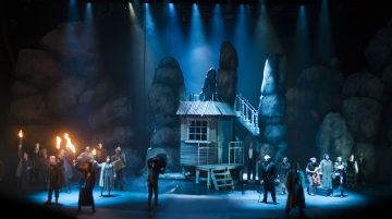 Teatterin näyttämölle on asetettu kalliomaisemat, pieni puinen hökkeli puujaloilla ja dramaattinen sininen valaistus. Useat aikakauden pukuihin pukeutuneet näyttelijät pitelevät soihtuja ja kokoontuvat ryhmiin, mikä luo jännittyneen ja dramaattisen tunnelman.