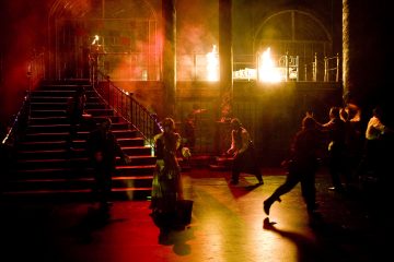 Näyttelijöitä hämärässä näyttämöllä, jossa on dramaattinen punainen valaistus, jotkut juoksevat ja toiset seisovat, kun kirkkaat liekit palavat taustalla portaikon ja pylväiden takana. Kohtaus vaikuttaa jännittyneeltä ja kaoottiselta.