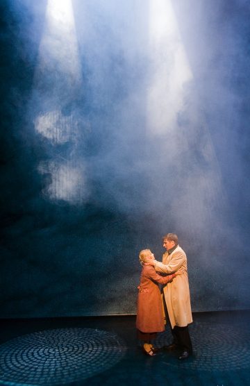 Kaksi pitkiin takkeihin pukeutunutta ihmistä syleilee dramaattisten valonsäteiden alla pimeällä, sumuisella näyttämöllä, mikä luo tunnelmallisen ja tunteikkaan tunnelman.