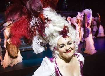 Lavalla hymyilee esiintyjä, jolla on hienostunut valkoinen peruukki, jossa on punaisia höyheniä, ja joka on pukeutunut yksityiskohtaiseen aikakauden pukuun. Muut esiintyjät, joilla on samanlaiset koristeelliset asusteet, hämärtyvät värikkäässä taustassa.