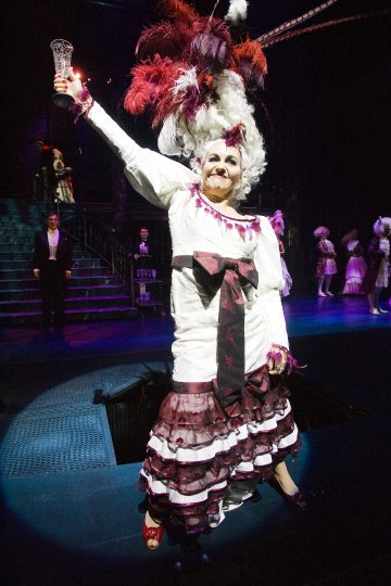 Valkoisen ja ruskeankeltaisen puvun ja suuren höyhenpäähineen omaava esiintyjä pitää lasia ylhäällä ja hymyilee lavalla muiden puvustettujen näyttelijöiden ja dramaattisen valaistuksen ympäröimällä näyttämöllä.