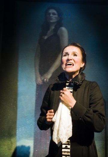 Tummaan pukuun ja röyhelökaulukseen pukeutunut nainen pitelee nenäliinaa ja vaikuttaa näyttämöllä tunteelliselta; hänen takanaan on heijastettu kuva pitkämekkoisesta naisesta, joka seisoo kädet ristissä ja katsoo eteenpäin.