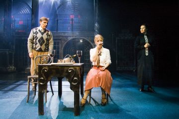 Lavalla esiintyy kolme näyttelijää; argyle-paitainen mies seisoo pöydän ääressä, jossa on shakkinappuloita ja viiniä, vaaleanpunaiseen hameeseen pukeutunut nainen istuu pitämällä kättään suullaan, ja toinen mustapukuinen nainen seisoo kädessään klarinetti.