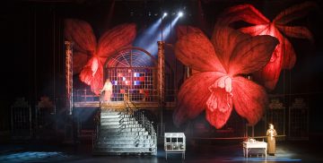 Dramaattisessa lavastuksessa on kaksi näyttelijää, suuret portaat, metalliportit ja ylimitoitetut punaiset kukkatarvikkeet tummalla taustalla, jota valaisee teatterivalo.