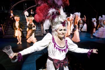 Lavalla poseeraa nainen, jolla on hienostunut valkoinen puku ja pitkä, höyhenpeitteinen peruukki, ja hänellä on lasi kädessään. Häntä ympäröivät muut esiintyjät, jotka ovat pukeutuneet koristeellisiin, 1700-luvun tyylisiin pukuihin dramaattisen näyttämövalaistuksen alla.