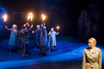 Joukko trenssitakkisia ihmisiä pitelee taskulamppuja ja elehtii vilkkaasti pimeällä näyttämöllä, kun taas vaalea takkiin pukeutunut nainen seisoo yksin etualalla ja katsoo poispäin ryhmästä.