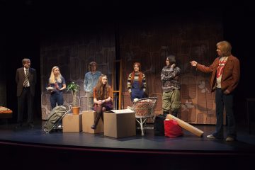 Kuusi ihmistä on näyttämöllä teatterikohtauksessa, jossa on liikkuvia laatikoita ja rekvisiittaa. Jotkut seisovat, jotkut istuvat, ja yksi henkilö elehtii muiden katsellessa. Taustalla on kuvioituja paneeleita, jotka viittaavat sisätiloihin.