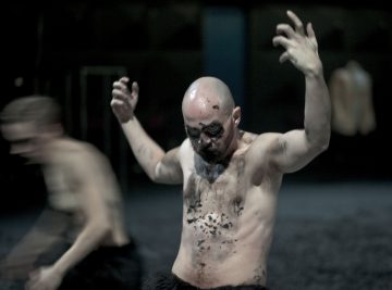 Paidaton mies, jolla on kalju pää ja mustat kasvot ja vartalomaali, nostaa dramaattisesti kätensä ylös, kun toinen epäselvä hahmo liikkuu hänen vieressään tummalla taustalla.