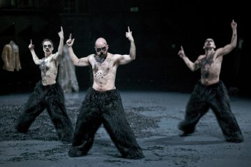 Kolme esiintyjää, joilla on maalatut kasvot ja tekoparrat, pukeutuvat mustiin karvaisiin housuihin ja tanssivat kohotetut etusormet kohotettuna hämärässä teatteriympäristössä. Lattia on täynnä tummia roskia.