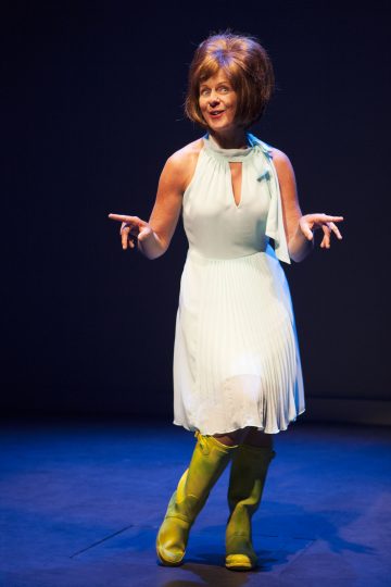 Nainen, jolla on hihaton valkoinen mekko ja kirkkaankeltaiset saappaat, seisoo lavalla ja elehtii elävästi molemmilla käsillään, valokeilassa tummaa taustaa vasten.