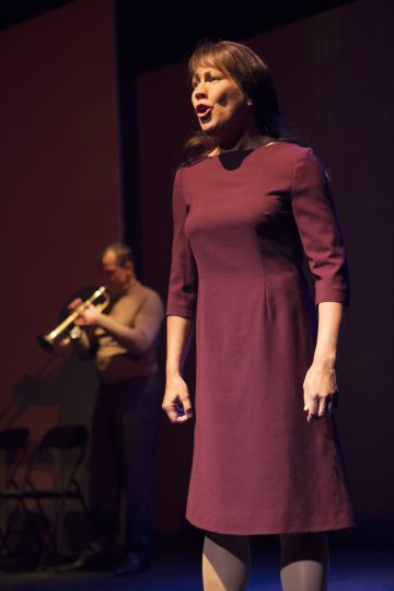 Lavalla laulaa viininpunaiseen mekkoon pukeutunut nainen, joka seisoo valokeilassa, kun mies taustalla soittaa trumpettia. Lavalla on tummat, vähäeleiset puitteet.