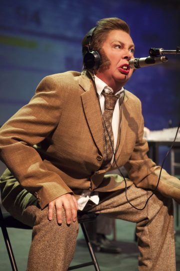 Ruskeaan pukuun pukeutunut henkilö istuu tuolilla, nojaa eteenpäin ja puhuu vilkkaasti mikrofoniin. Hänellä on kuulokkeet ja näyttämömeikki, liioitellut kasvonpiirteet ja partapiikit, jotka viittaavat esitykseen tai teatteriesitykseen.