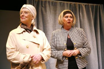 Kaksi vanhempaa naista seisoo harmaan verhon edessä. Toisella on kermanvärinen takki ja päähuivi, ja hänellä on kädessään turkisstola. Toisella on yllätetyn näköinen mustavalkoinen sudenhattutakki ja musta päähine.