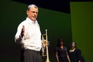 Valkoiseen paitaan pukeutunut mies pitelee lavalla trumpettia ja elehtii toisella kädellään, ja taustalla seisoo kaksi mustiin mekkoihin pukeutunutta naista vihreää taustaa vasten.