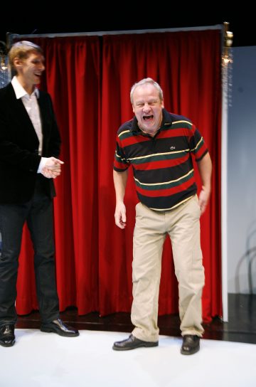 Kaksi miestä seisoo punaisen verhon edessä. Toinen mies, jolla on raidallinen poolopaita ja khakihousut, nauraa sydämellisesti. Toinen mies, jolla on tumma bleiseri ja valkoinen paita, hymyilee ja katsoo häntä kohti.