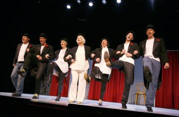 Seitsemän mustiin takkeihin, valkoisiin paitoihin ja mustiin hattuihin pukeutunutta ihmistä esittää lavalla koreografioidun korkean potkun rutiinin hymyillen, taustalla punaiset verhot ja lavavalot.